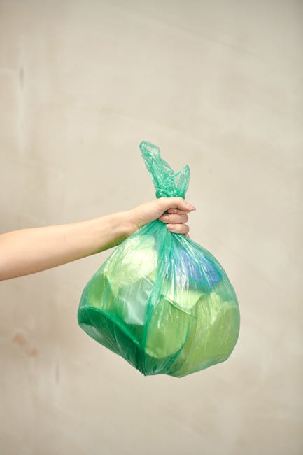 A person's hand and forearm extend from the left side of the image, holding a green plastic rubbish bag tied at the top with a knot, against a plain, light-colored background. The bag appears partiall