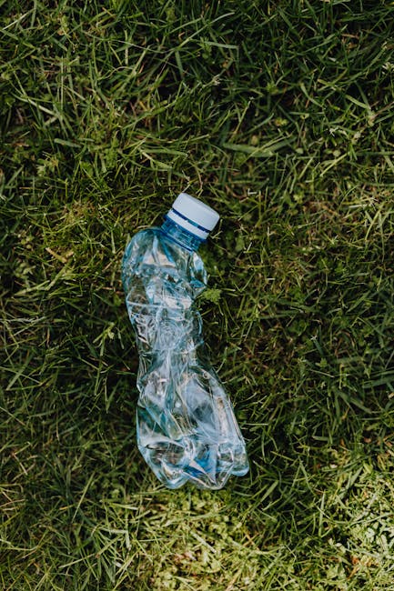 A person's hand and forearm extend from the left side of the image, holding a green plastic rubbish bag tied at the top with a knot, against a plain, light-colored background. The bag appears partiall