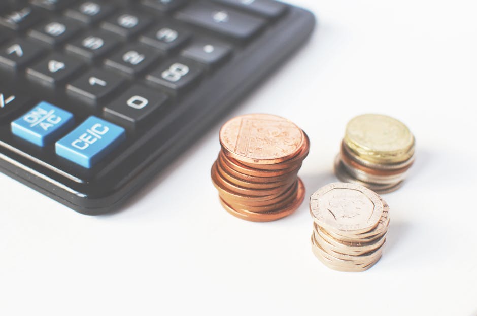 A close-up image displays a black calculator with blue and white keys positioned in the upper left corner, alongside three neatly stacked piles of coins on a white surface. The first pile, situated in