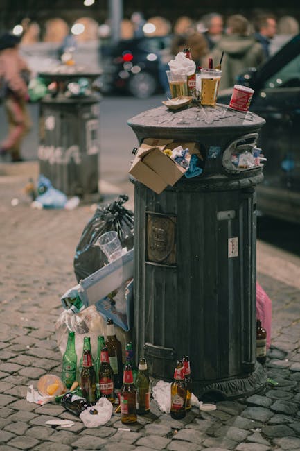 A black, cylindrical public trash bin made of metal with a domed top, situated on a cobblestone sidewalk in an urban environment. The bin's surface shows signs of wear, with graffiti and accumulated d