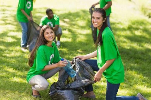 A group of young volunteers, wearing matching bright green T-shirts with a recycling symbol, are participating in a community cleanup in a grassy outdoor area. Two women are prominently positioned in the foreground, smiling at the camera while holding a large black rubbish bag together; one is crouching and holding a plastic water bottle near the bag, and the other is kneeling, grasping the bag's opening. In the background, two other volunteers, also dressed in green T-shirts, are seen beside a second rubbish bag and appear to be involved in collecting debris. The scene is set in a well-lit, sunny environment with lush green grass and trees, emphasizing a positive and environmentally conscious atmosphere. This image visually highlights community efforts in rubbish collection, aligning with professional waste management services such as those offered by House Clearance Pimlico, serving local areas including Pimlico and the surrounding London regions.